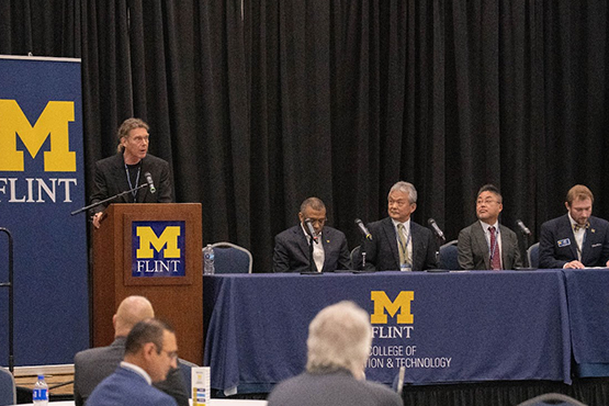 A speaker at a podium addresses an audience during an event at the University of Michigan-Flint, with a panel of speakers seated behind.