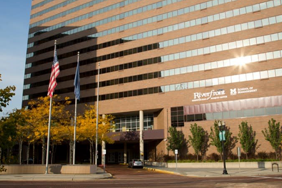 The exterior of the Riverfront building, featuring flags, trees with yellow leaves, and the School of Management sign.