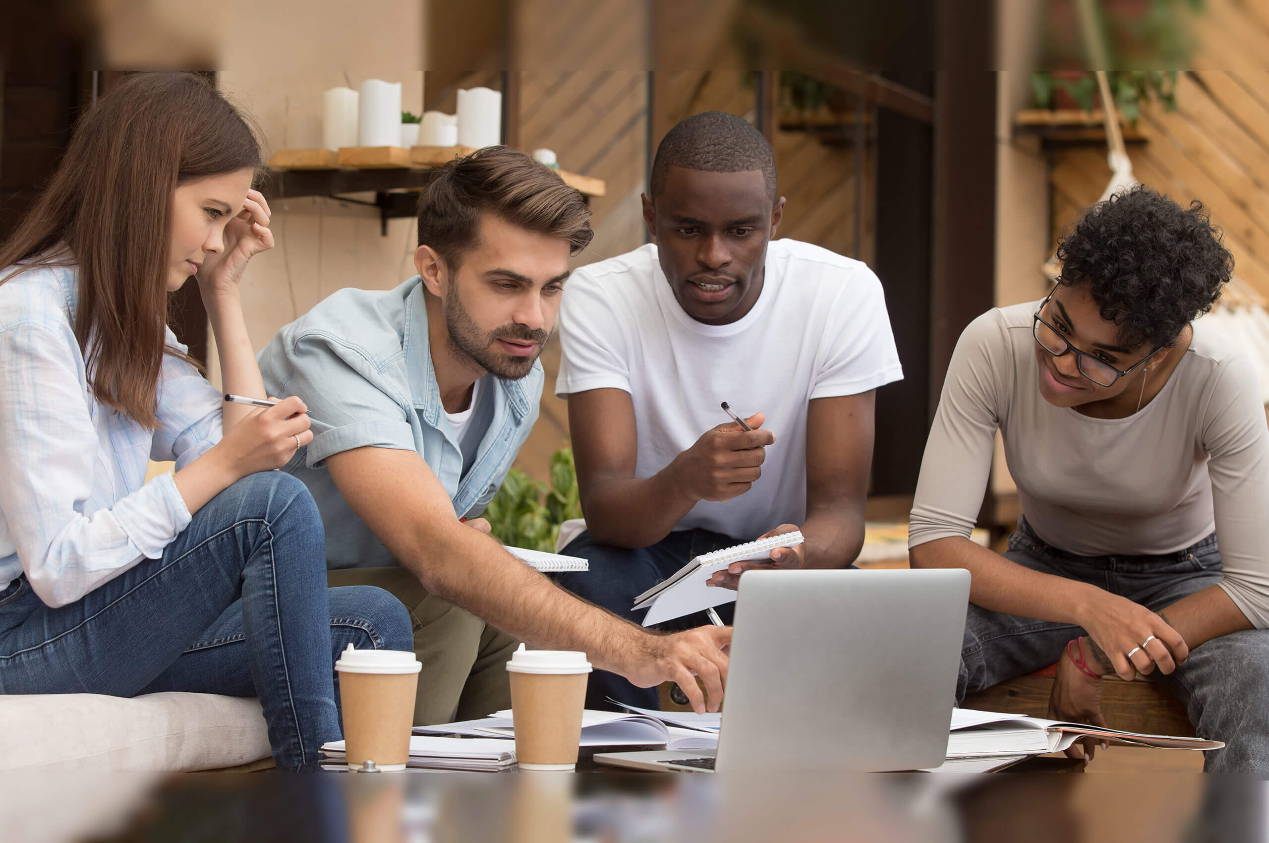 A group of five people is gathered around a laptop, discussing and taking notes. There are coffee cups and papers on the table.