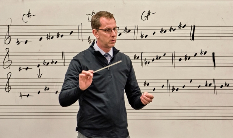 A person holding a conducting baton in front of a whiteboard with musical notes and symbols, including C# and G#.