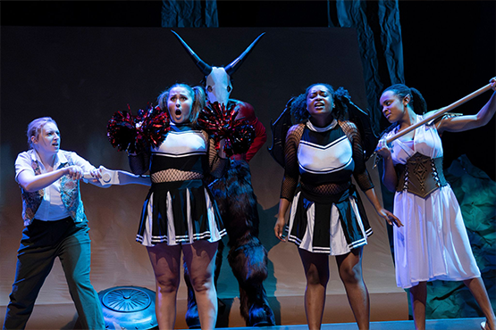 A theatrical scene with four actors and a figure wearing a goat mask in dramatic lighting.