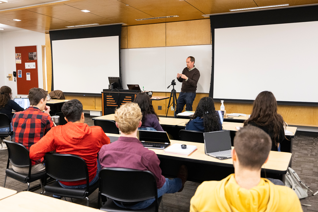 A classroom setting with a speaker presenting in front of two large screens. Students are seated at desks, some with laptops open, attentively listening.