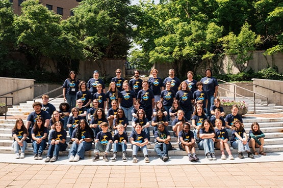 A large group of diverse children and adults wearing matching blue t-shirts with a logo, posing on outdoor steps surrounded by greenery.