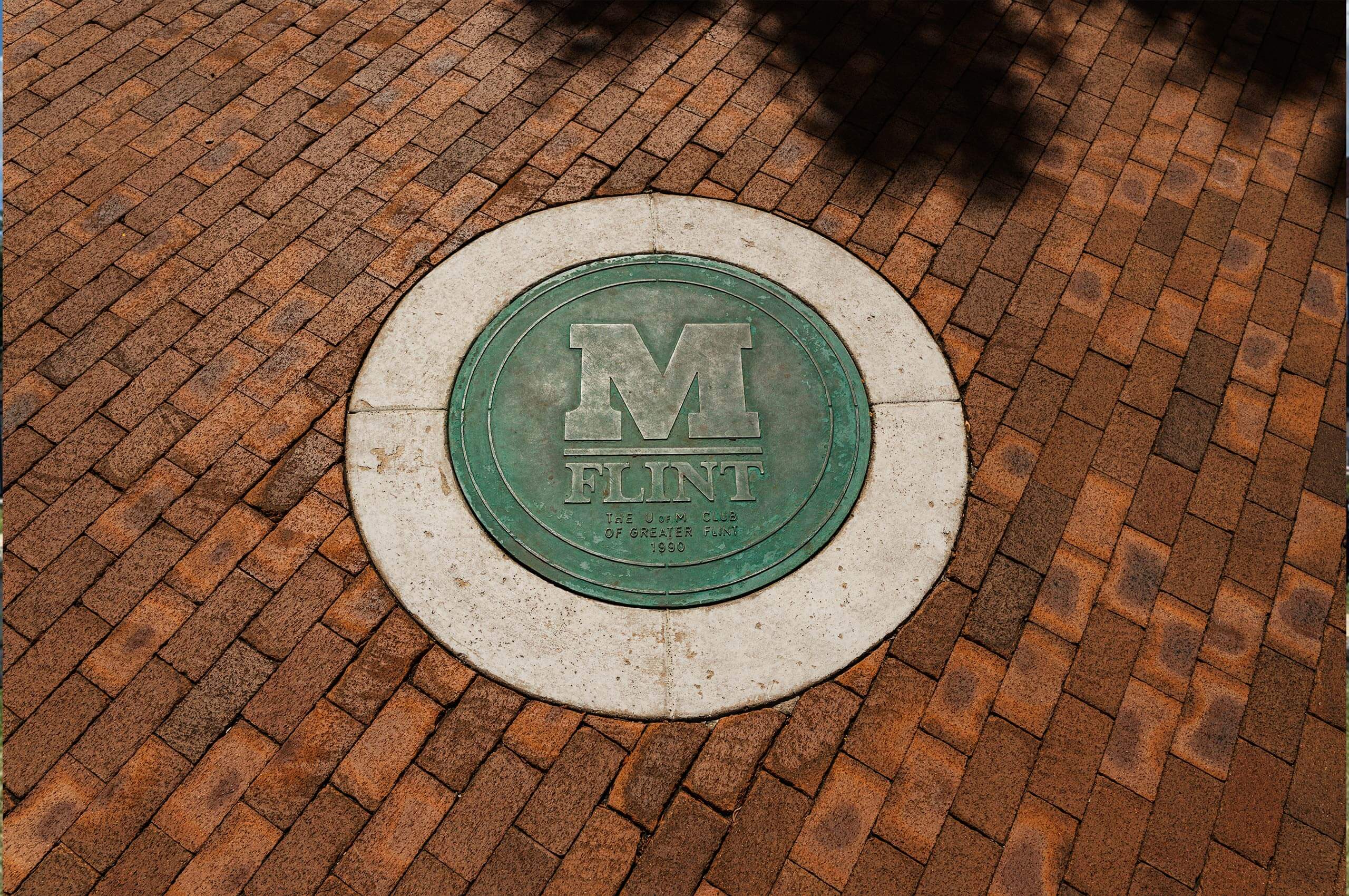 A circular bronze plaque embedded in a brick walkway, featuring the letter "M" and the word "FLINT," with the text "THE JAYCEES OF GREATER FLINT 1960."