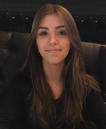 Giulia Angelina Colone with long, wavy hair is seated in a dark, stylish environment, wearing a black top and a delicate necklace.