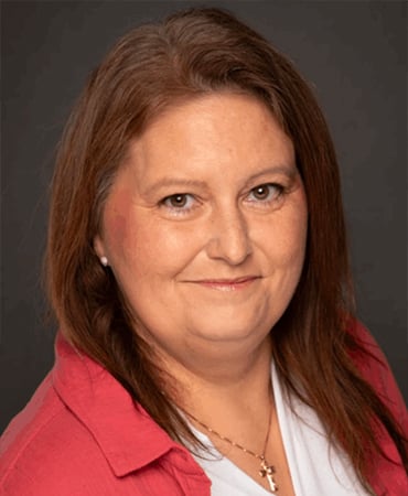 Laura Lynne Lucas with long brown hair is wearing a red jacket over a white top, with a necklace visible. The background is dark.