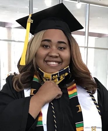 Mya Baxter wearing a graduation cap and gown, adorned with colorful stoles, poses for a photo indoors.