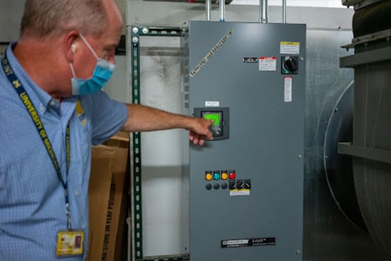 A person wearing a blue shirt and a University of Michigan lanyard points at a control panel with buttons and a small screen in an industrial setting.