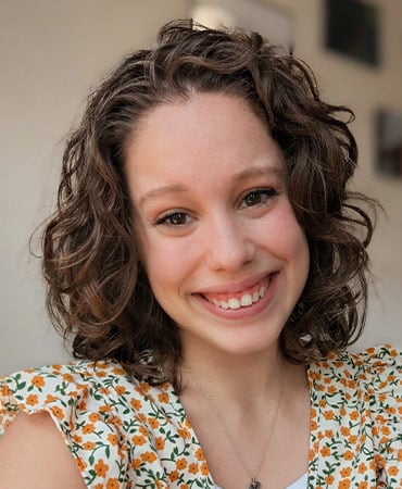 Emma C. with curly brown hair wearing a floral patterned top and a necklace.