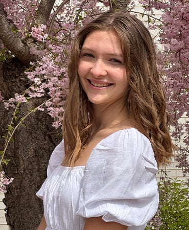 Kaitlyn with long wavy hair wearing a white puff-sleeve blouse standing in front of a tree with pink blossoms.