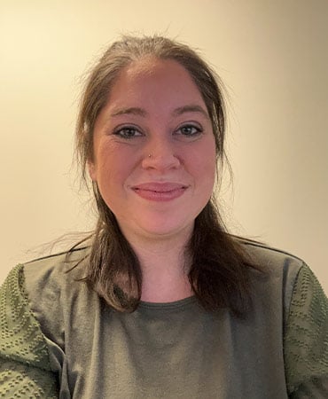 MacKenzie with shoulder-length brown hair wearing an olive green shirt with textured sleeves against a plain beige background.