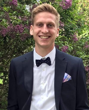 Larry Popyk wearing a dark suit jacket, white shirt, and a black bow tie with white polka dots, standing outdoors in front of green foliage and purple flowers.