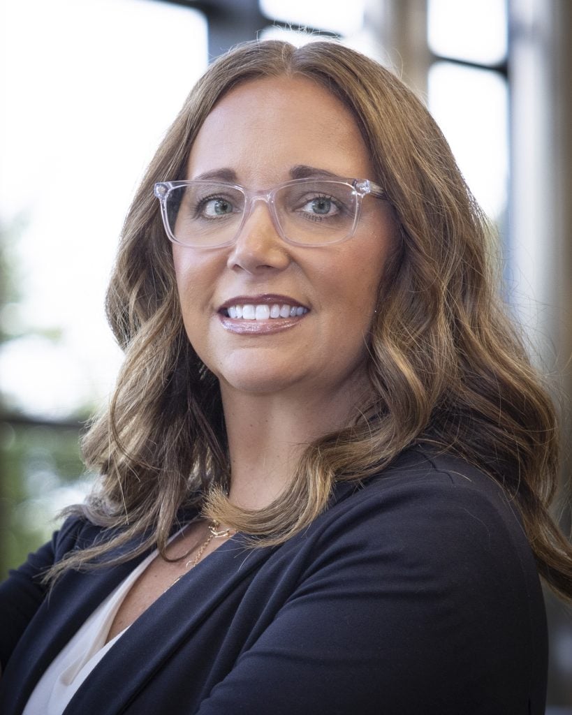 Suzanne Adams with long wavy hair wearing glasses, a dark blazer, and a white top, standing indoors near windows.