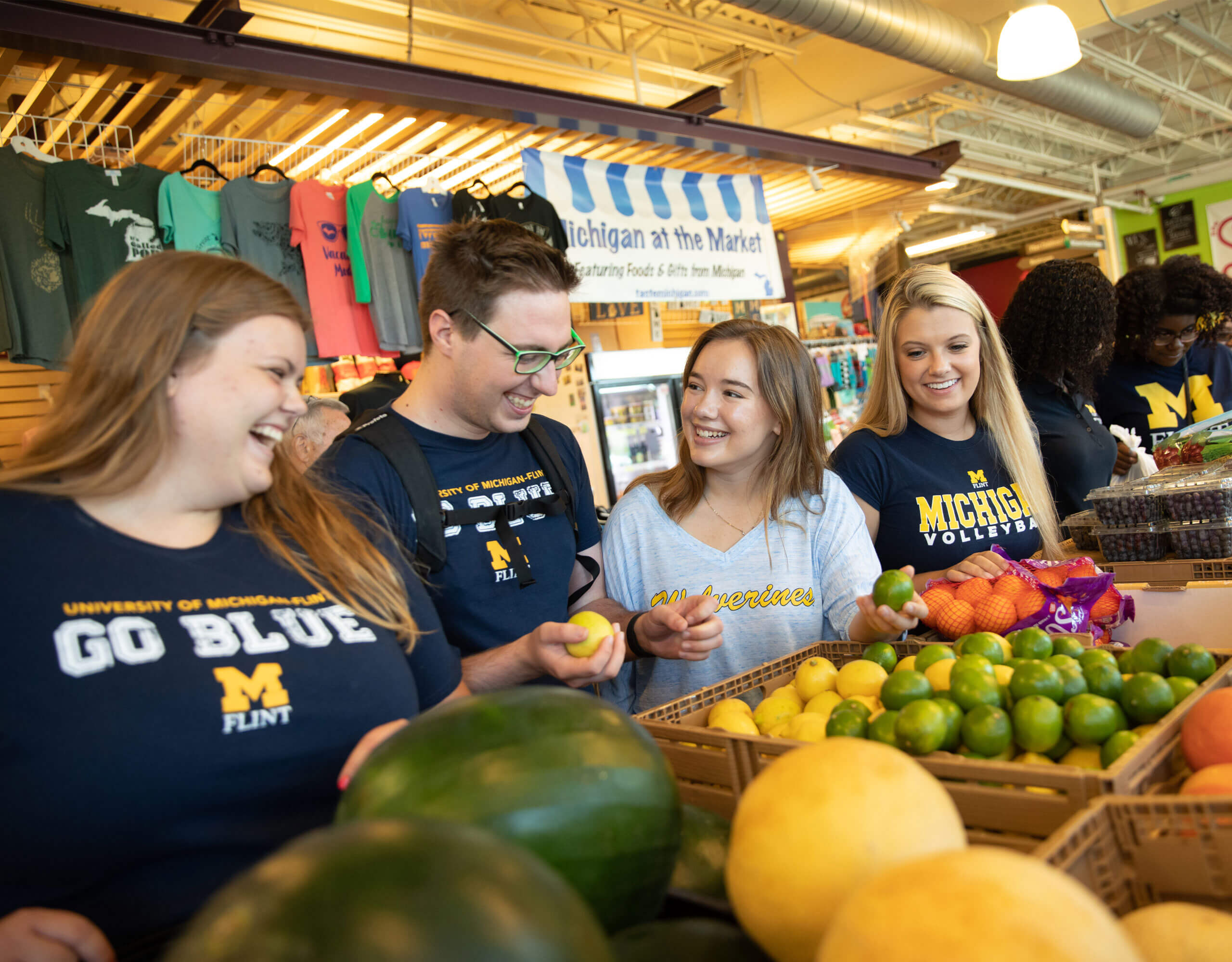 A group of people in University of Michigan apparel stands at a market stall filled with various fruits and vegetables.