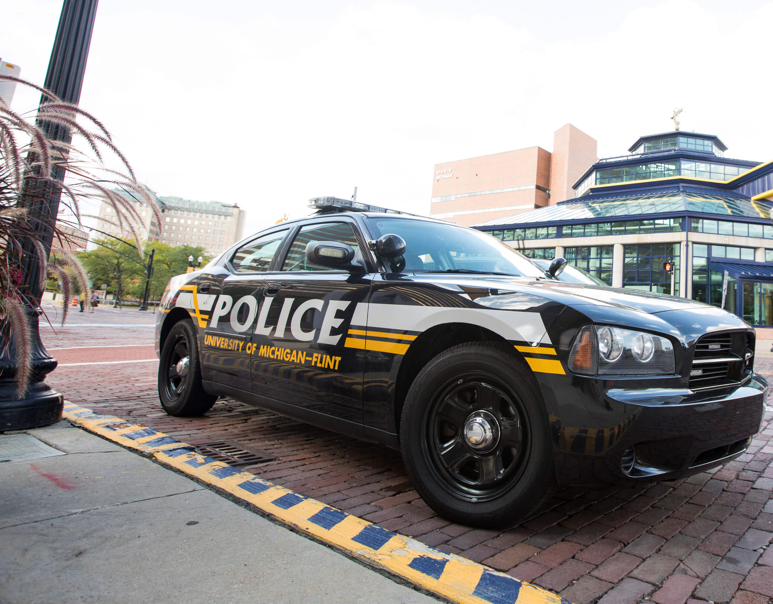 A University of Michigan-Flint police car parked on a brick street, with a modern building in the background.