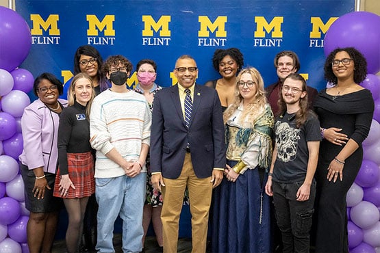 A group of people posing together in front of a blue backdrop with the letters "M" and "FLINT" in yellow, surrounded by purple balloons.
