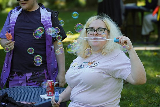 A person blowing bubbles with a bubble wand, surrounded by floating bubbles, while holding a can of soda on a table.