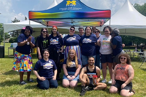 A group of diverse individuals poses together in front of a colorful tent that reads "CENTER FOR GENDER AND SEXUALITY." They are outdoors on a sunny day, with green grass and white tents in the background.