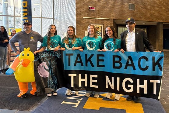 A group of five people holding a large banner that reads "TAKE BACK THE NIGHT," with one person in a duck costume and another in a suit.