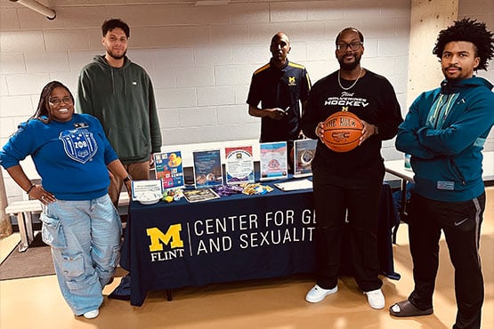 A group of four people standing behind a table displaying materials from the University of Michigan Flint's Center for Gender and Sexuality. One person is holding a basketball.