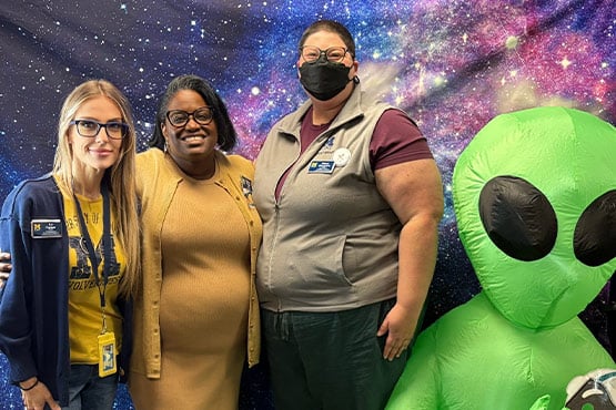 Three women pose together in front of a colorful galaxy backdrop, with a large green alien figure beside them.