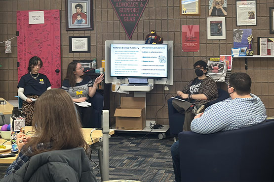 A group of four people seated in a room with a presentation screen displaying text. Two individuals are speaking while the others listen. The room has various posters and decorations on the walls.