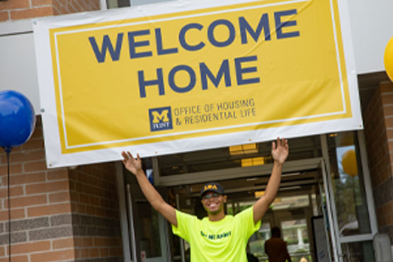 Residente de habitação segurando placa Welcome Home na entrada da First Street Residence Hall em UM-Flint.