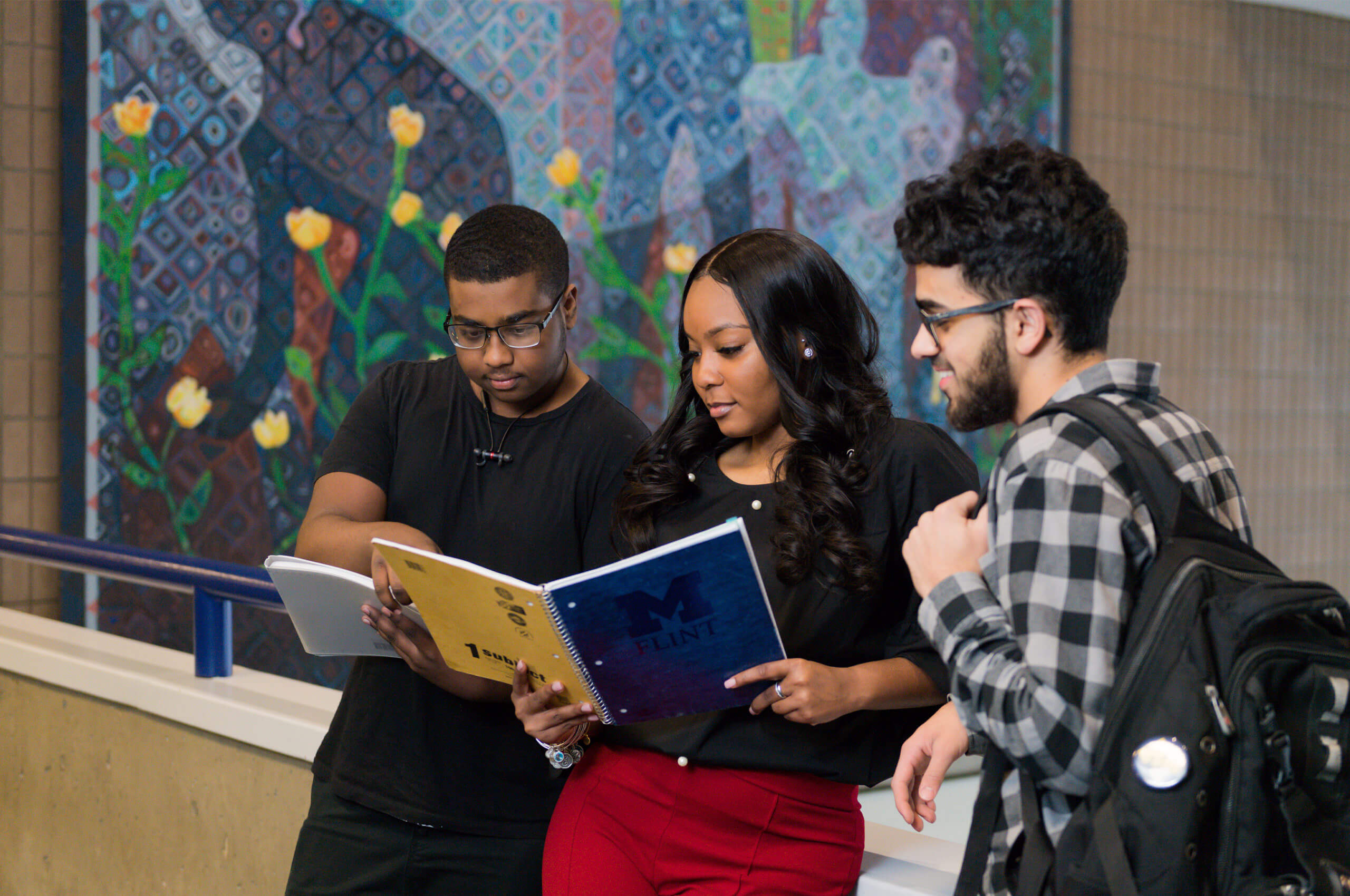 Three students are gathered around a notebook, discussing its contents, with a colorful mural in the background.