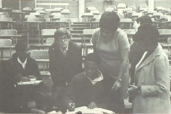 A group of five people in a classroom with empty desks, engaged in discussion or study.