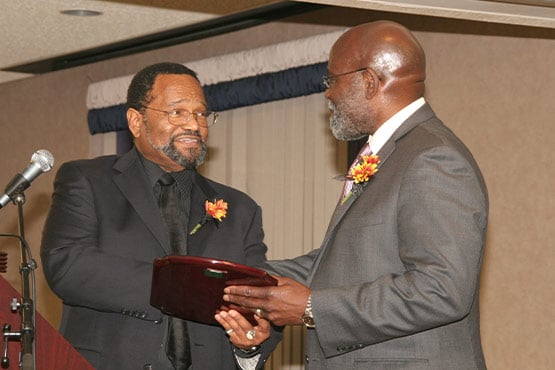 Two men in suits exchanging a plaque or award at an indoor event.