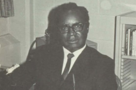 A black and white photo of a person in a suit and tie sitting at a desk in an office with bookshelves and a computer.