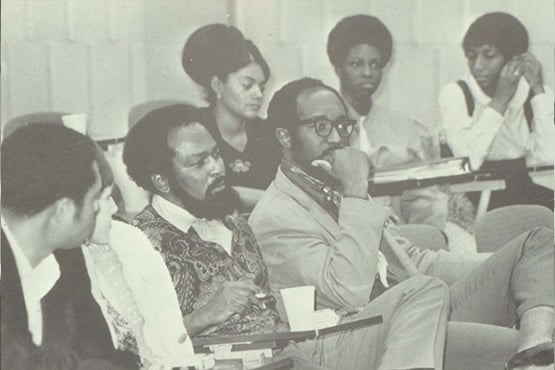 Black and white photo of six people sitting in a classroom or lecture hall, engaged in discussion or listening.