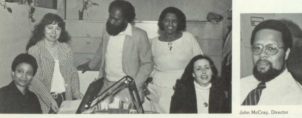 A group of five people in an office setting, with filing cabinets and a desk lamp visible. Next to this is a photo of John McCray.