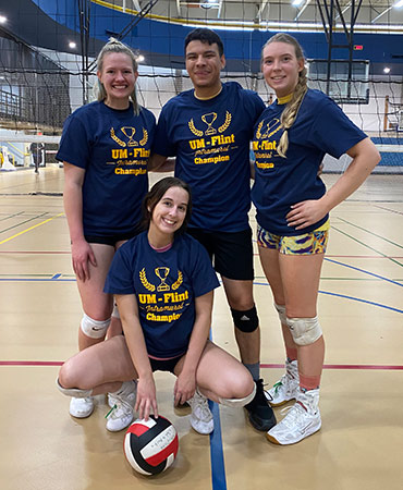Four volleyball players pose together in a gym, wearing matching blue shirts that say 