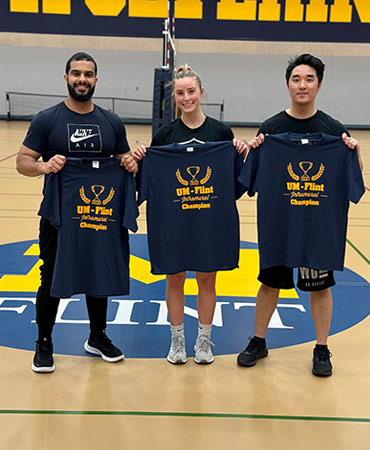 Three individuals stand on a gym floor holding navy blue t-shirts that read 