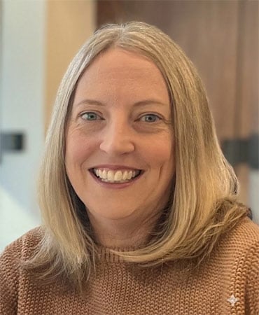 Tamra Watt with long, straight blonde hair wearing a brown sweater, sitting indoors with a blurred background.