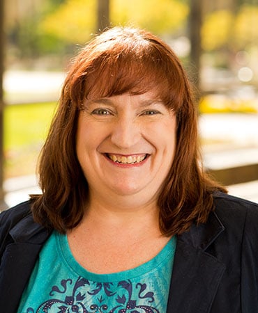Anna Reed smiles for a professional headshot. She is wearing a green shirt that has a black design with a black suit jacket overtop. Her medium-length, red hair is hanging onto her shoulders with bangs hanging overtop her forehead.