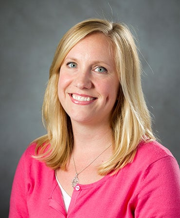 Rachel Gutierrez smiles for a professional headshot. She is wearing a pink, buttoned sweater and a silver necklace with a pendant. Her medium-length, blonde hair is hanging down onto her shoulders.