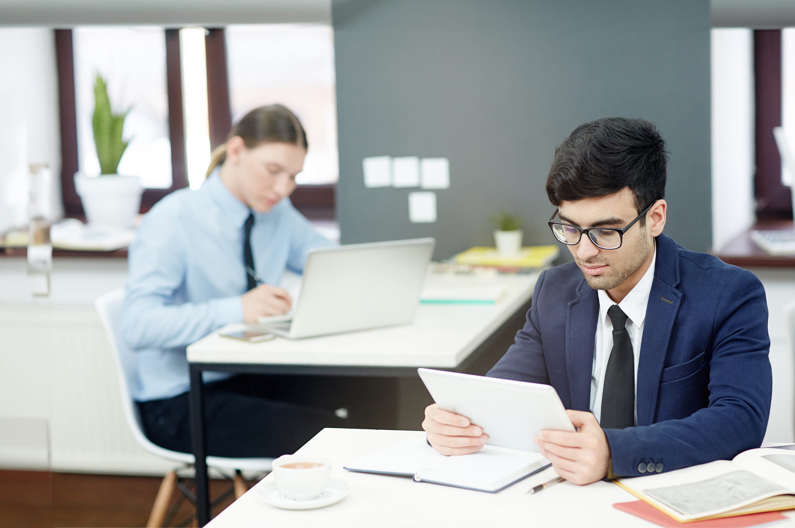 A man in a suit holds a tablet while sitting at a table in a modern office, with another person working on a laptop in the background.