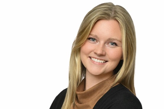 Person with long blonde hair wearing a brown turtleneck and a black sweater against a white background.