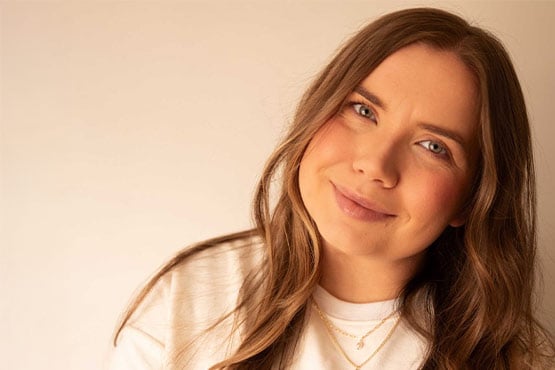 Person with long brown hair wearing a white top and layered gold necklaces against a beige background.