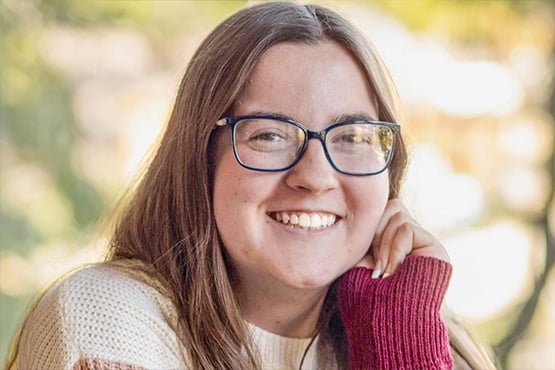 Emma Swanson with long brown hair wearing glasses and a white sweater with red sleeves, resting their chin on their hand.