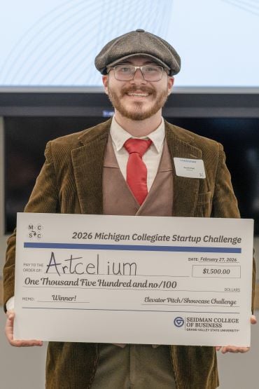 Austin Hall wearing a brown corduroy jacket, red tie, and flat cap holding a large check for ,500 made out to "Artcelium" as the winner of the 2026 Michigan Collegiate Startup Challenge.
