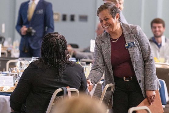 A person in a gray blazer is greeting another person seated at a dining table in a formal setting.