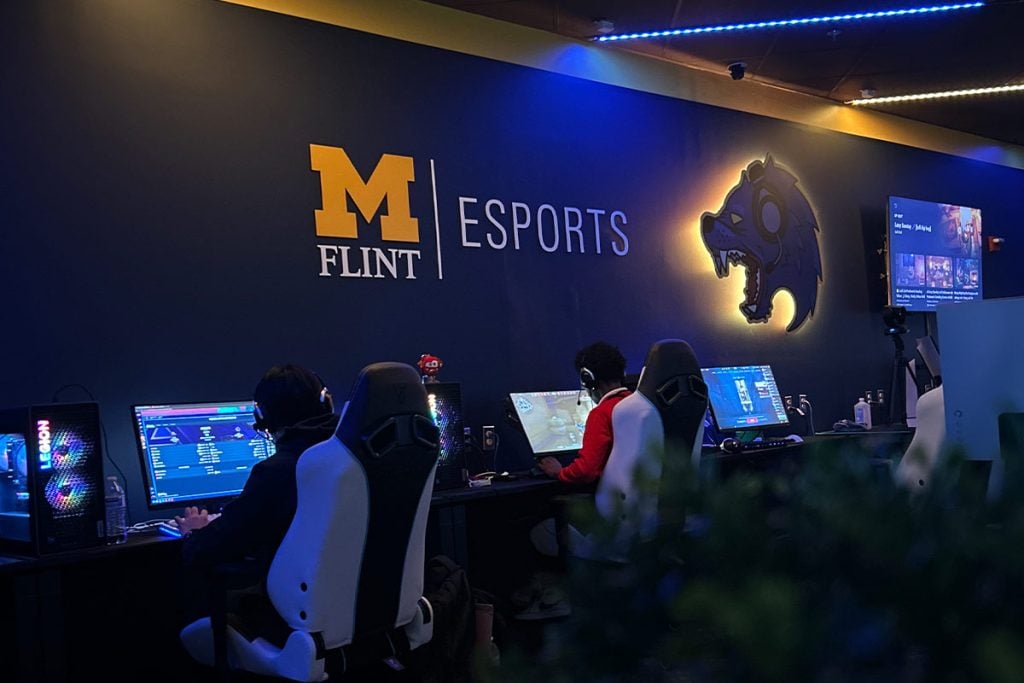 A gaming setup featuring two players at computers in a dimly lit esports arena, with a wall displaying "M FLINT ESPORTS" and a wolf logo.