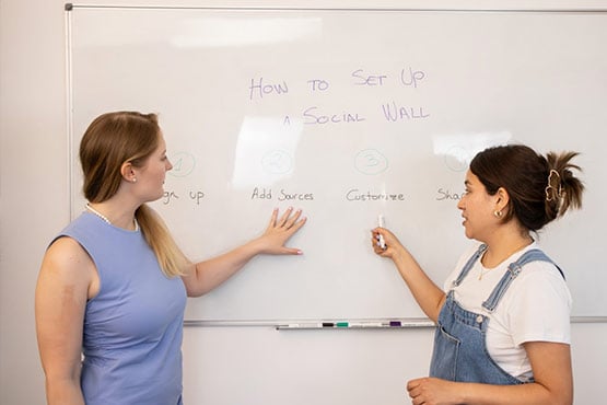 Two individuals are standing in front of a whiteboard with the text "How to Set Up a Social Wall" and four steps listed: "Set Up," "Add Sources," "Customize," and "Skim." One person is pointing at the board while the other holds a marker.