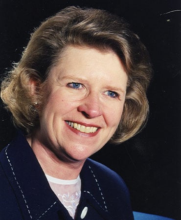 Beverly Schmoll with short, wavy blonde hair wearing a dark blue jacket with white stitching and a white blouse underneath.
