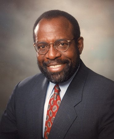 Charlie Nelms wearing a dark suit, white shirt, and red patterned tie against a neutral studio background.