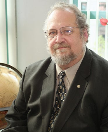 Jack Kay wearing a dark suit and patterned tie sitting indoors near a globe and window.