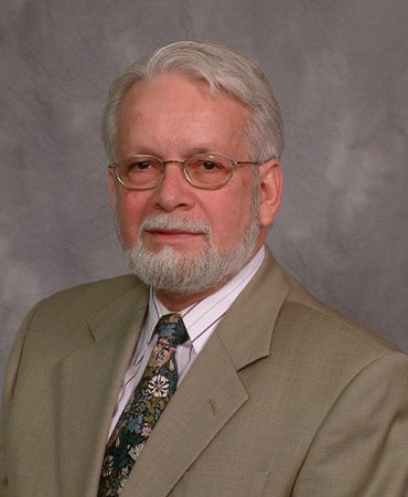 Juan Mestas with gray hair wearing a tan suit jacket, white shirt, and floral patterned tie against a gray background.